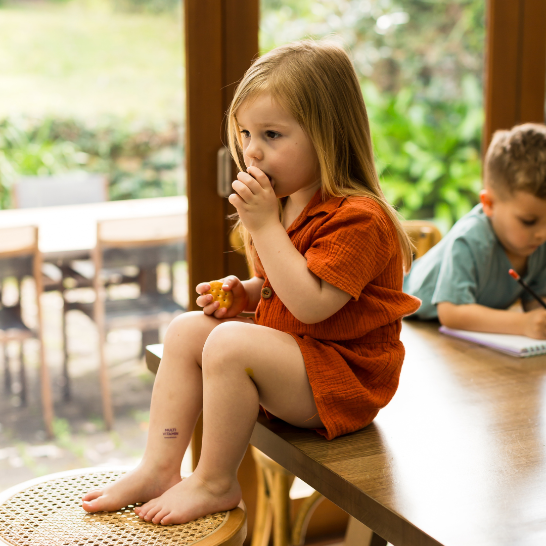 Young child sitting on a table enjoying a snack while another child is drawing - both wearing multivitamin patches